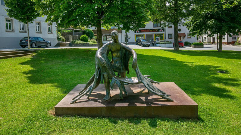 Schutzengel für Neuhausen: Eine Bronzestatue mit organischen Formen steht auf einer grünen Wiese unter Bäumen, im Hintergrund sind ein Auto und Gebäude zu sehen.