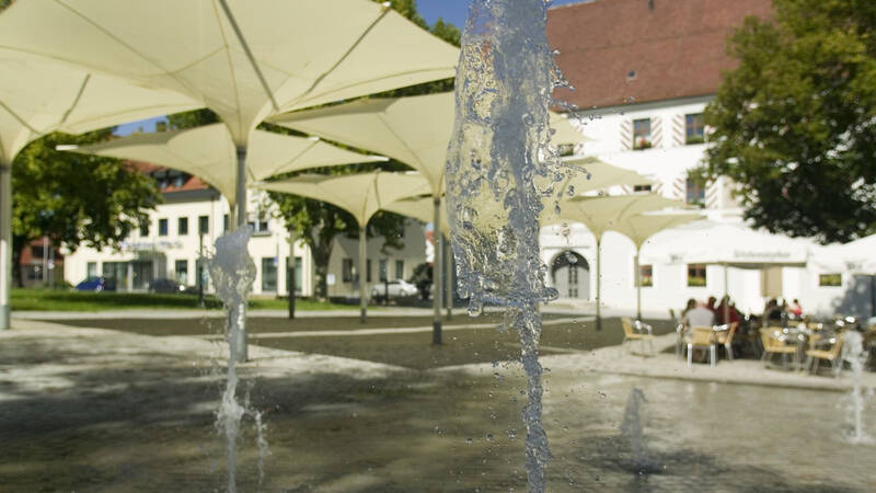 Ein Platz mit Wasserspielen im Vordergrund, überdacht von modernen Segeltuchkonstruktionen, und im Hintergrund ein Gebäude mit Menschen, die an Tischen im Freien sitzen.