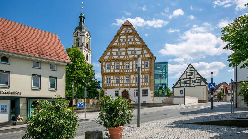 Das Bild zeigt eine malerische Altstadt-Szene mit Fachwerkhäusern, einer Kirche mit Turm und einem klaren, blauen Himmel.