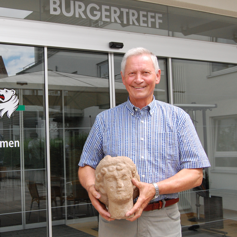 Der Archäologische Weg: Ein lächelnder älterer Mann steht vor dem Eingang eines Bürgertreffs und hält eine Steinbüste in den Händen.