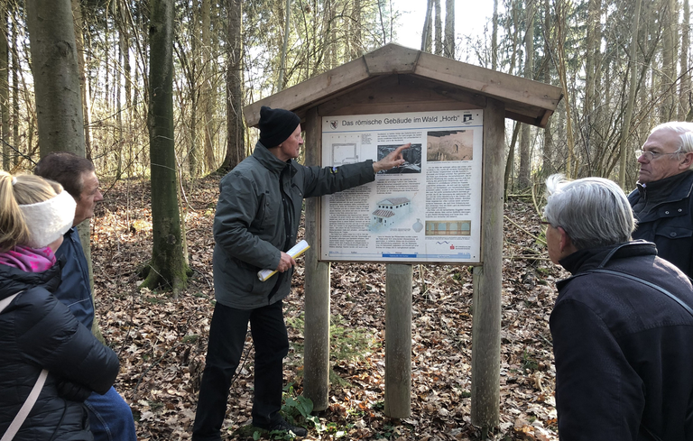 Der Archäologische Weg im Sauhag und Horber Wald: Eine Gruppe von Personen steht im Wald um ein Hinweisschild, während ein Mann Informationen darauf zeigt und erklärt.