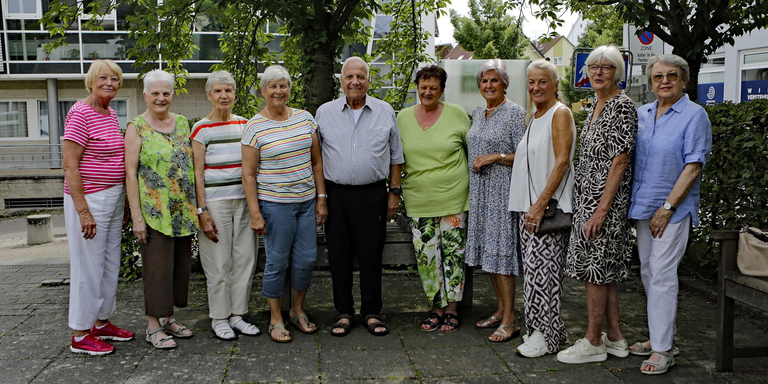 Das Team des Besuchsdienstes: Eine Gruppe älterer Menschen steht lächelnd im Freien in einer Reihe, umgeben von Bäumen und Gebäuden.