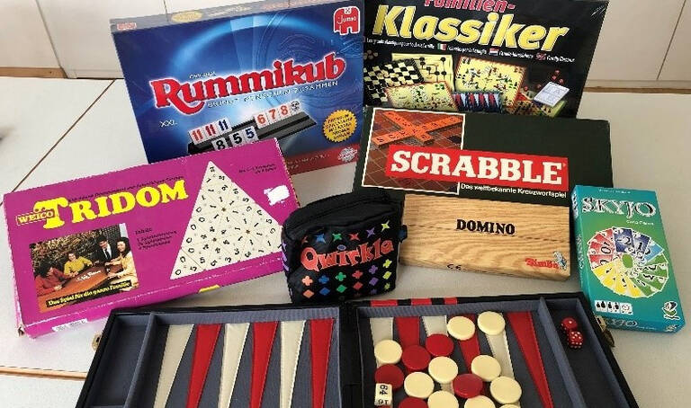 Spieletreff im Bürgertreff iAuf dem Bild sind verschiedene Gesellschaftsspiele wie Rummikub, Scrabble, Tridom, Quirkle, Domino und Skyo zu sehen, die auf einem Tisch ausgebreitet sind.