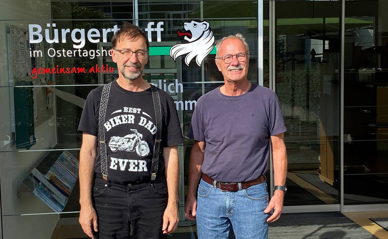 Für Sie da - Rat und Tat: Zwei Männer stehen lächelnd vor einem Gebäude mit der Aufschrift "Bürgertreff im Ostertagshof".