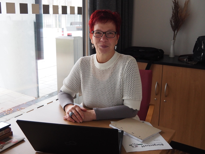 Eine Person mit rotem Haar und Brille sitzt an einem Tisch mit einem Laptop und Papieren vor einem großen Fenster.