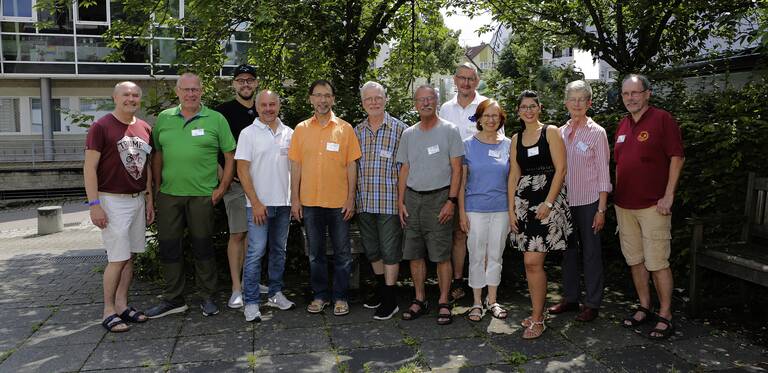 Mitarbeitende Reparaturcafé - Eine Gruppe von zwölf Personen steht draußen auf einem gepflasterten Weg vor einem Gebäude und Bäumen.